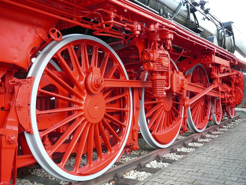 Close-up view of the red wheels and mechanical components of a steam locomotive. The large spoked wheels and detailed parts are visible, showcasing the intricate design and engineering of the historic train—a marvel from an era without today's government dilemmas. The locomotive is situated on a track.