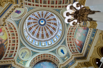 The ornate, detailed dome ceiling of the grand government building is decorated with intricate murals, gold accents, and architectural embellishments. A large chandelier with frosted lamps hangs from the ceiling, surrounded by vibrant paintings and blue-and-gold designs.