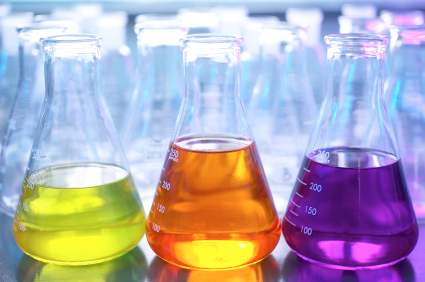 Three conical flasks, filled with yellow, orange, and purple liquids respectively, are neatly arranged in a row on a laboratory bench. With various other out-of-focus lab glassware in the background, they form the perfect setup for an engaging chemistry lesson.