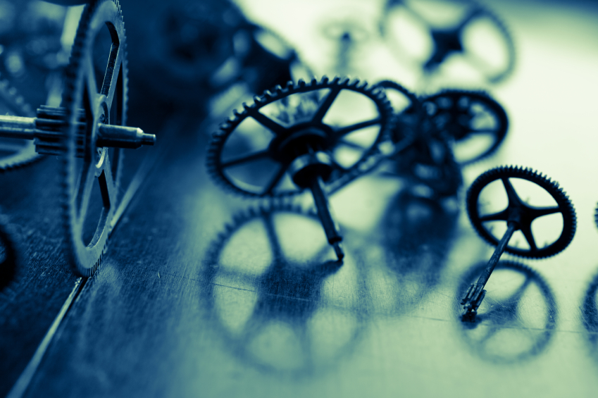 Close-up of several interconnected gears and cogs scattered on a dark surface. The image has a blue tint and a shallow depth of field, highlighting the intricate details and metallic texture of the components, symbolizing the precision required in preparing tomorrow's leaders.