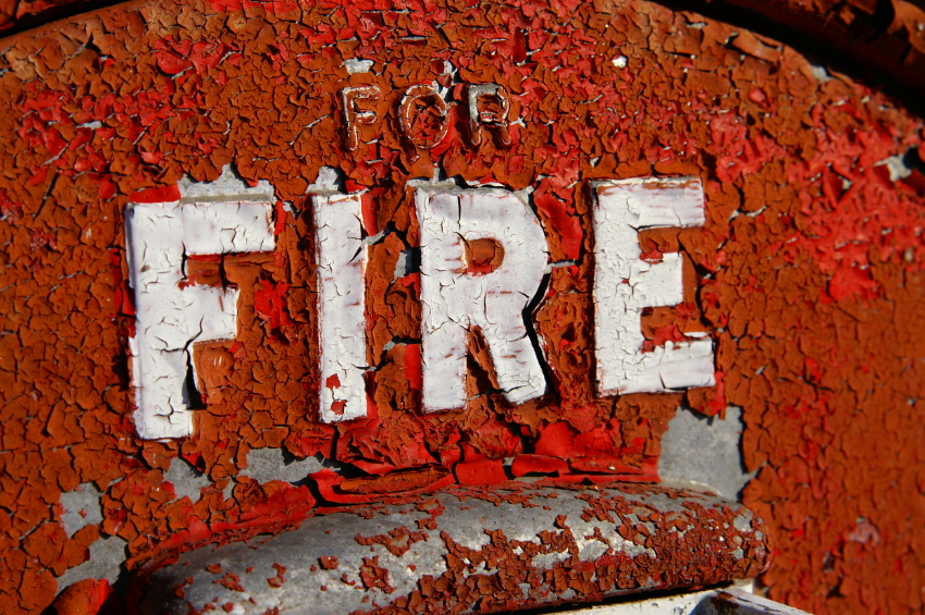 Close-up of a rusted and peeling metal surface with the word "FIRE" in large white letters. The surrounding area is painted red, but the paint is chipped and cracked, revealing the metal underneath. The text "FOR" appears above "FIRE" in smaller letters, reminiscent of old emergency services signage.