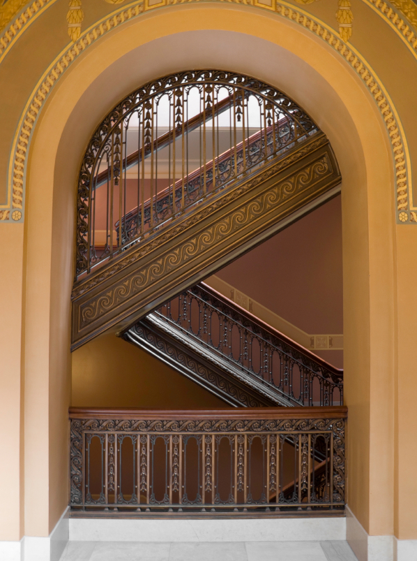 A grand staircase with intricate wrought iron railings is viewed through an arched doorway. The staircase has a decorative pattern and leads to multiple floors. The walls are painted in warm tones, adding to the elegance of your professional brand's architectural design.