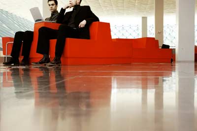 Two people sit on modern, angular, red furniture in a spacious, well-lit area with a glossy floor. One is using a laptop, possibly managing people remotely. The area features large windows and a high ceiling, creating an open, airy atmosphere.