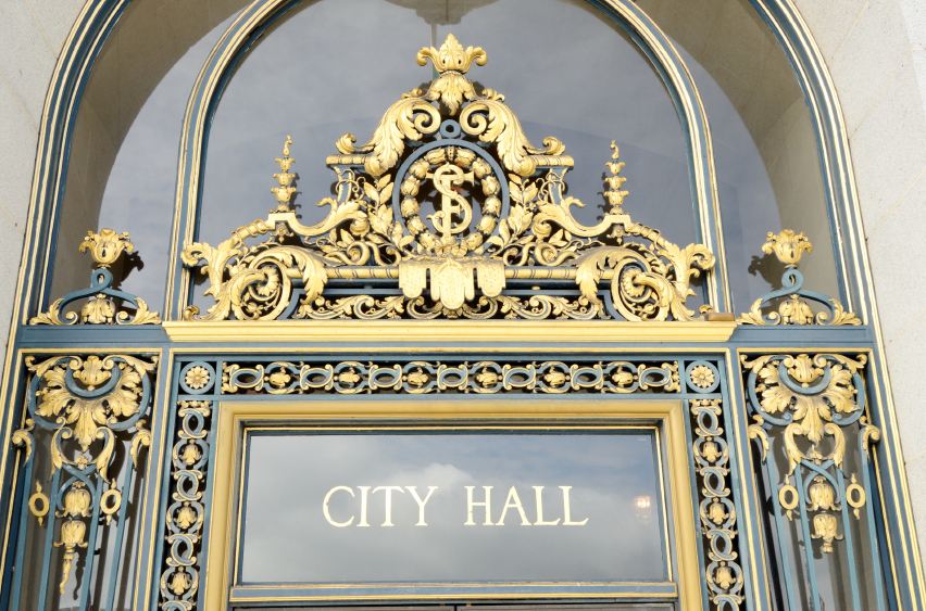 An ornate entrance to City Hall featuring detailed gold decorations and a prominent circular insignia at the top center. Emphasizing the integrity of public service, the words "CITY HALL" are displayed on a glass panel below the intricate designs. The archway is made of elaborate ironwork and stone.