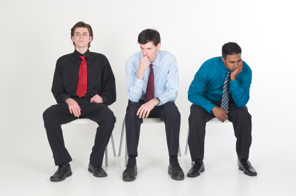 Three men in business attire sit on chairs against a white background, appearing deep in thought as if they are interviewing. The man on the left wears a black shirt with a red tie, the center man wears a light blue shirt with a maroon tie, and the man on the right wears a teal shirt with a striped tie.