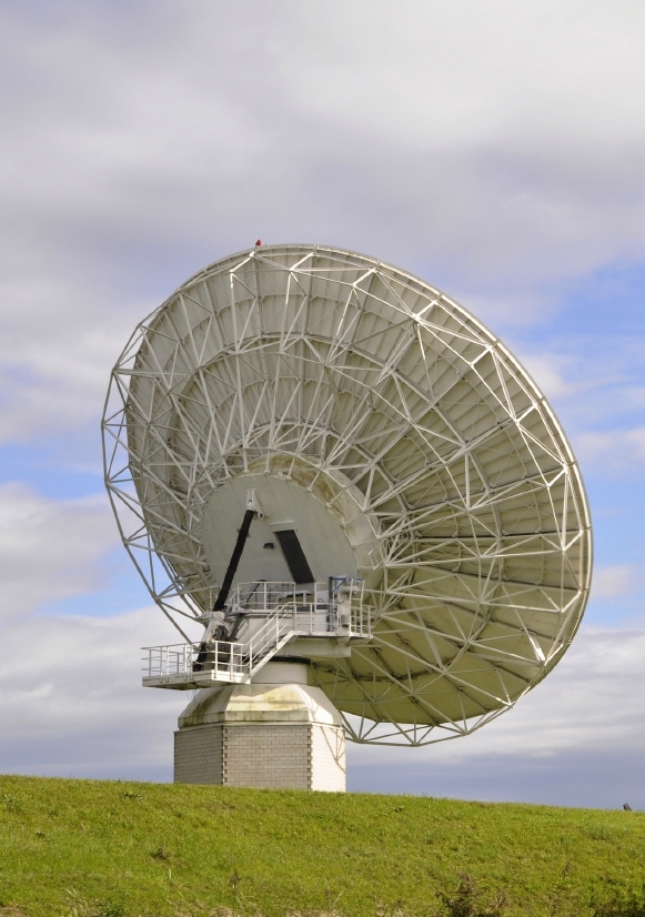 A large satellite dish made of metal with a white and gray color scheme is positioned on a grassy hill. The sky in the background is mostly cloudy, with a few patches of blue visible. The dish, essential for networking, is angled slightly upward and mounted on a concrete base.