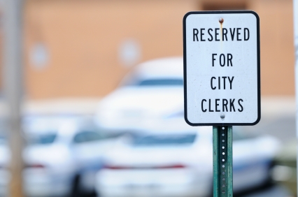 A sign reads "Reserved for City Clerks" in black text on a white background. The sign is mounted on a metal post, with blurred cars and a building visible in the background, symbolizing the essential roles in the public sector.