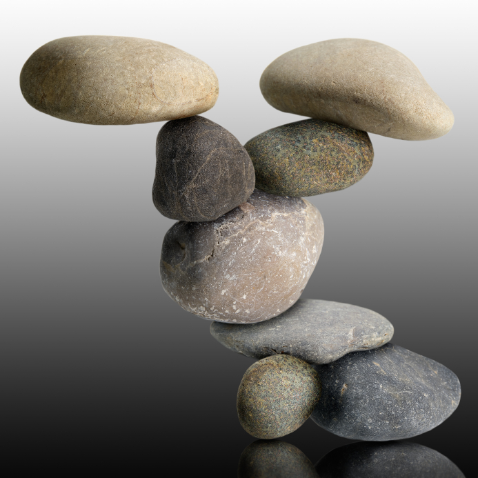 A balanced stack of variously shaped and colored smooth stones. The stones are arranged in a gravity-defying formation, symbolizing the equilibrium of balancing career and personal life. The background transitions from white at the top to dark gray at the bottom.