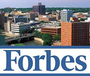 A colorful aerial view of a city with various mid-sized buildings juxtaposed against a backdrop of greenery and a river. The prominent word "Forbes" is displayed in large blue text across the lower part of the image, hinting at its recognition as a top career website.