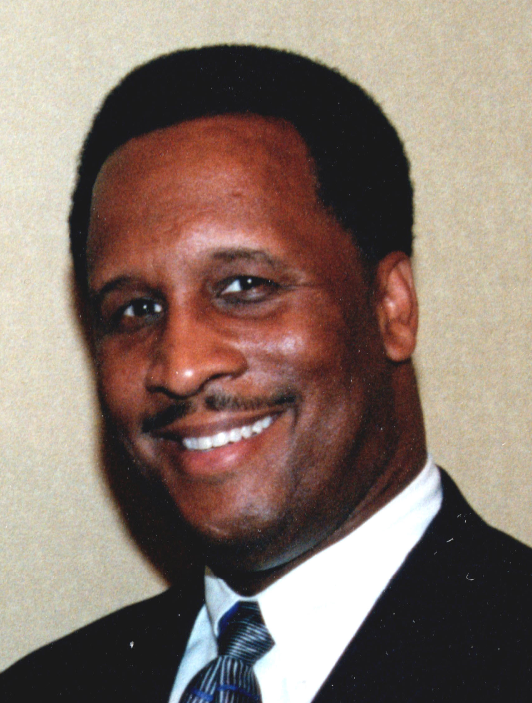 A man with short curly hair, wearing a dark suit, white shirt, and patterned tie, is smiling at the camera, exuding leadership. The background is a plain light-colored wall.