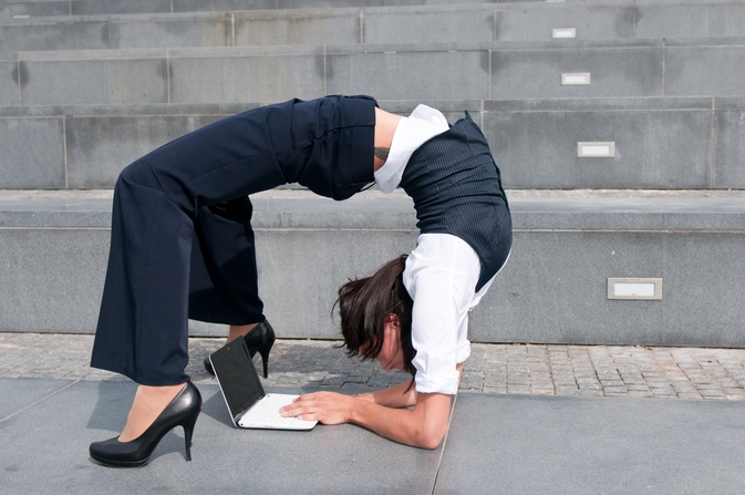 A person dressed in business attire, including high heels and a white shirt, performs an impressive backbend while working on a laptop on the ground. The background features gray concrete steps.