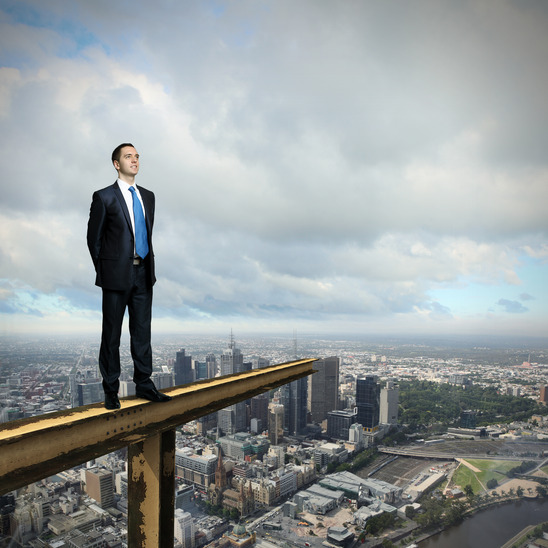 A man in a suit and tie stands confidently at the edge of a high metal beam, overlooking a vast cityscape below with buildings, streets, and greenery. The sky is cloudy, creating a dramatic backdrop as he contemplates his next job.