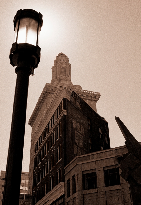 A sepia-toned image of a tall, historic government building with an intricate tower, viewed from below. A lamppost stands prominently in the foreground, and other urban structures are partially visible around the main building.