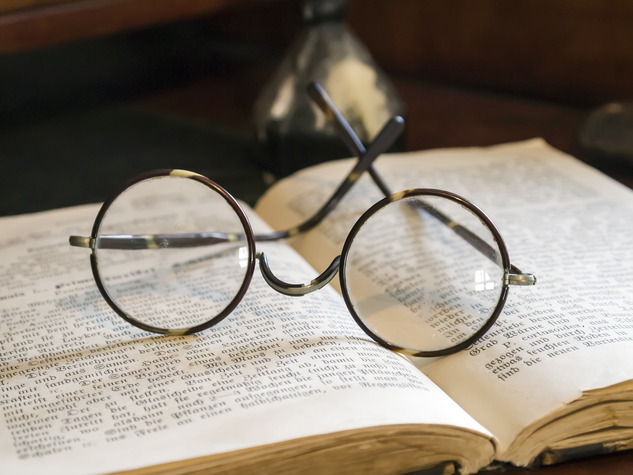 A pair of vintage round eyeglasses with wire frames rests open on an old book with faint text. The background features a blurred, dark object, possibly a decorative item or tool, creating a warm, nostalgic atmosphere that evokes memories of a long, dedicated career.
