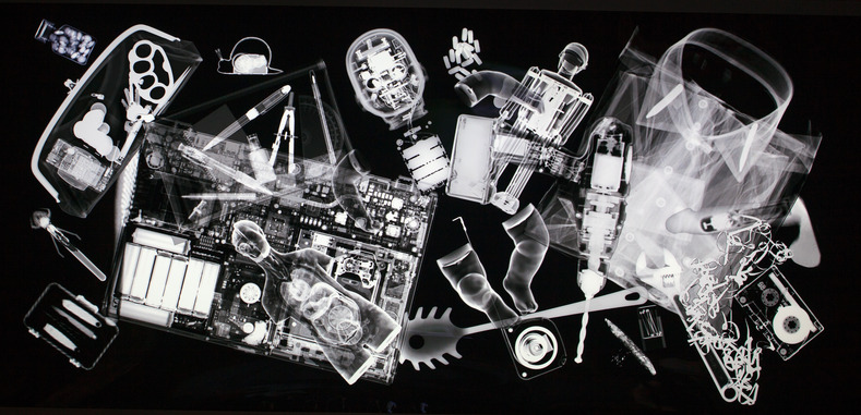 An X-ray image shows various objects including electronic circuits, a toy robot, tools, a shirt collar, kitchen utensils, screws, batteries, and assorted small items scattered across a dark background—all discoverable through an online archive.