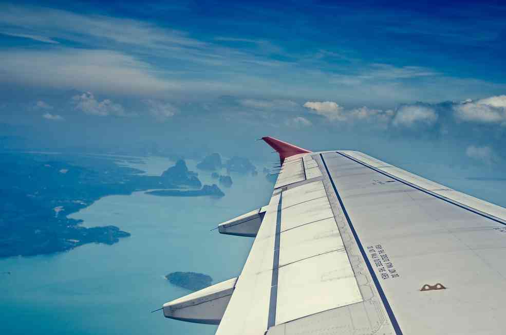 A view from an airplane window showing the aircraft's wing and engine, with a vast expanse of blue sky above and a scenic landscape of islands and water below. The horizon is visible in the distance, separating the sky from the land and sea, like borders set by nature's own government.