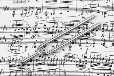 A tuning fork lies on top of a sheet of music. The sheet music is densely covered with various musical notes and symbols. The tuning fork, a metallic instrument with two prongs, is centrally placed on the sheet, underscoring its essential role in facilitating accurate pitches for public agencies and musicians alike.