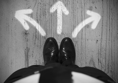 Black and white image taken from a first-person perspective, showing a pair of formal shoes standing on a concrete surface with three arrows painted on the ground, pointing left, right, and forward. The arrows sequester different paths, indicating a crucial choice of direction.