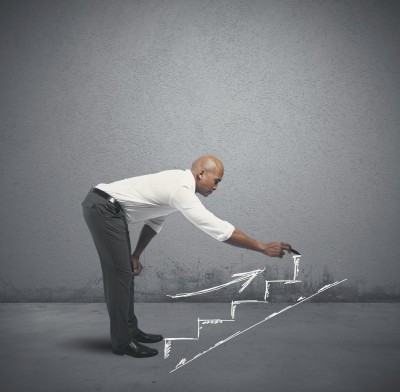 A man in business attire is drawing a staircase with chalk on a grey wall. Bending down to sketch the lower steps, he includes an upward arrow symbolizing growth and progress, much like submitting an application for personal or professional advancement. The scene vividly depicts a journey toward success.