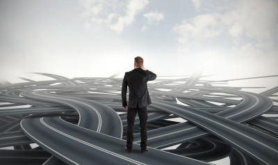 A man in a suit stands with his back to the camera, scratching his head in confusion while facing an intricate network of intertwined roads that extend into the cloudy sky. The image conveys a sense of uncertainty and complexity, much like choosing the best career for your major.
