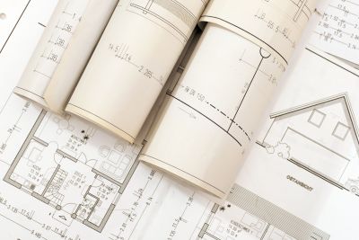 A stack of architectural plans and blueprints, including detailed floor layouts and house elevation drawings. Some of the plans are rolled up, while others are spread out, showcasing intricate measurements and design notes essential for a building project that aims to drive economic development.