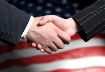 Two individuals in business attire shaking hands against a blurred background of the American flag, symbolizing successful public-private partnerships.