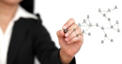 A person in a black suit draws a network diagram on a transparent surface. The diagram consists of small human icons connected by lines, illustrating the concept of networking and relationships among people. The background is white.