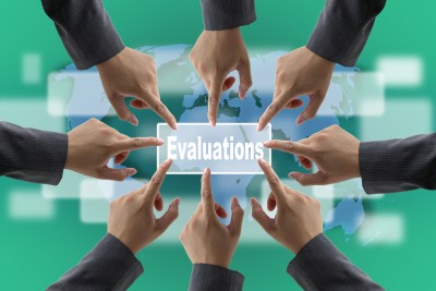 A graphic showing several hands in business suits pointing towards a central rectangular button labeled "Evaluations" against a world map background. The image suggests a collaborative focus on performance evaluation and appraisals.