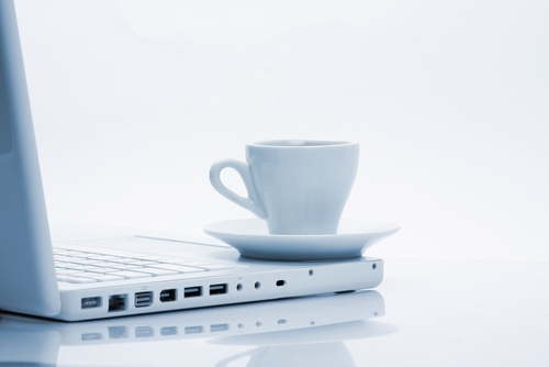 A white coffee cup on a saucer rests on a white surface next to a white laptop, seamlessly capturing the essence of teleworking. The image features a clean, minimalistic aesthetic with a light color palette, showcasing the laptop’s ports and part of the keyboard.
