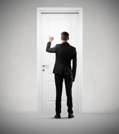 A person in a black suit is shown from behind, knocking on a white door. The individual, likely heading to an interview, has short brown hair and stands in a minimalist, gray-toned room with no visible decorations.