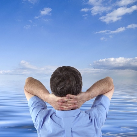 A person stands with their back facing the camera, hands clasped behind their head, gazing at a calm sea under a bright blue sky with scattered clouds. The scene evokes a sense of career contemplation and tranquility.