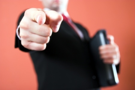 A person in a suit, holding a folder in their left hand, points directly at the camera with their right index finger, as if interviewing you. The background is a solid orange color, and the person's face is out of focus.