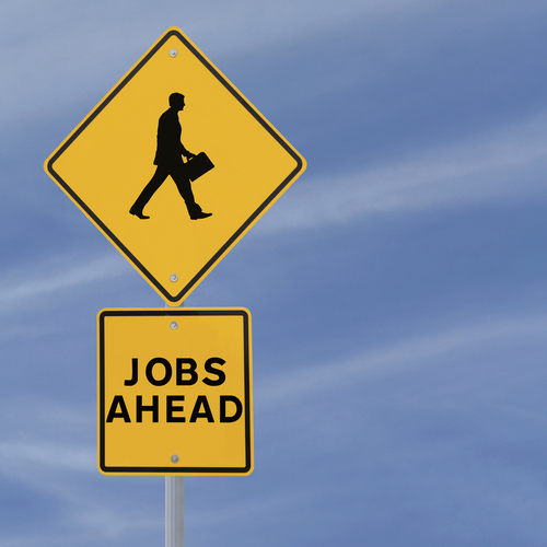 A yellow diamond-shaped road sign features the silhouette of a walking person carrying a briefcase. Below it, a rectangular yellow sign reads "JOBS AHEAD" in bold black letters. The word "Job" stands out prominently against the clear blue sky with faint wispy clouds.