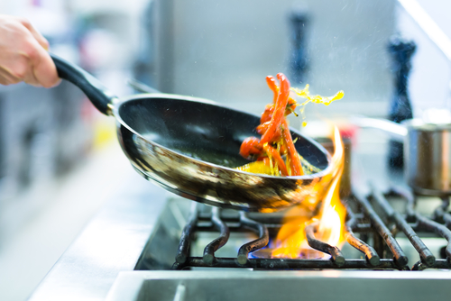 A person is demonstrating leadership by expertly sautéing vegetables in a black frying pan over a gas stove with visible flames. The vegetables, including red peppers and green herbs, are mid-air as the pan is tilted. The background shows a blurred kitchen setting.