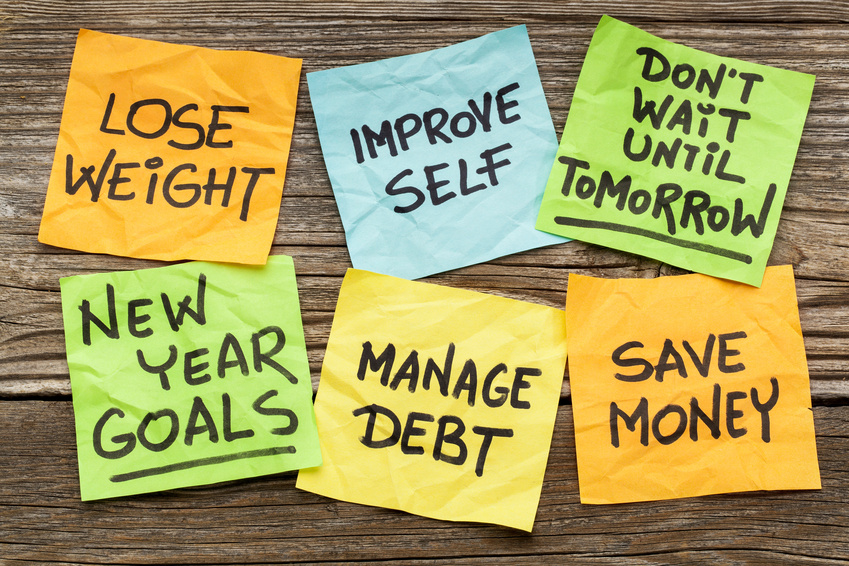 Six colorful sticky notes on a wooden surface display motivational messages for your resolutions: "LOSE WEIGHT," "IMPROVE SELF," "DON'T WAIT UNTIL TOMORROW," "NEW YEAR GOALS," "MANAGE DEBT," and "SAVE MONEY.