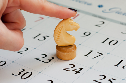 A hand with black nail polish gently touches a beige knight chess piece placed on a calendar. The dates 15, 23, and 24 are prominently visible, suggesting an emphasis on those days—perhaps pivotal moments in one's career. The background is slightly blurred, focusing attention on the hand and the chess piece.