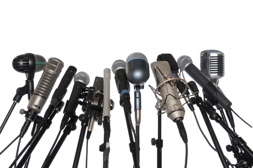 A collection of various types of microphones, including dynamic, condenser, ribbon, and handheld models, is arranged on adjustable microphone stands against a white background. The mics are positioned close together in a semi-circle formation, perfect for public speaking engagements.