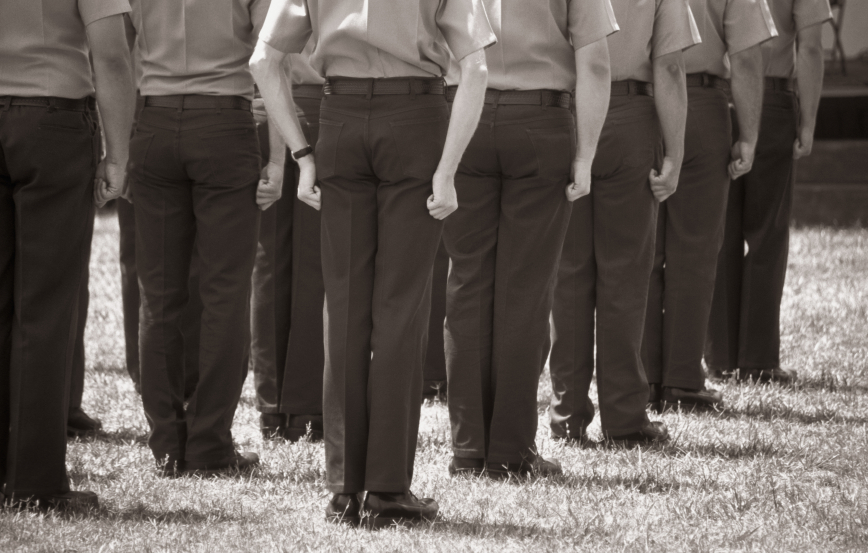 A sepia-toned image of job seekers standing in formation, seen from behind. They are wearing uniforms with hands clenched into fists hanging by their sides, and are standing on a grassy field.