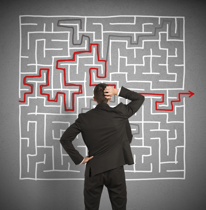 A person in a suit, seen from behind, stands in front of a complex maze drawn on a gray wall. The maze has a red path guiding from the entrance to the exit. The individual, likely contemplating their career path, scratches their head as they ponder the solution.