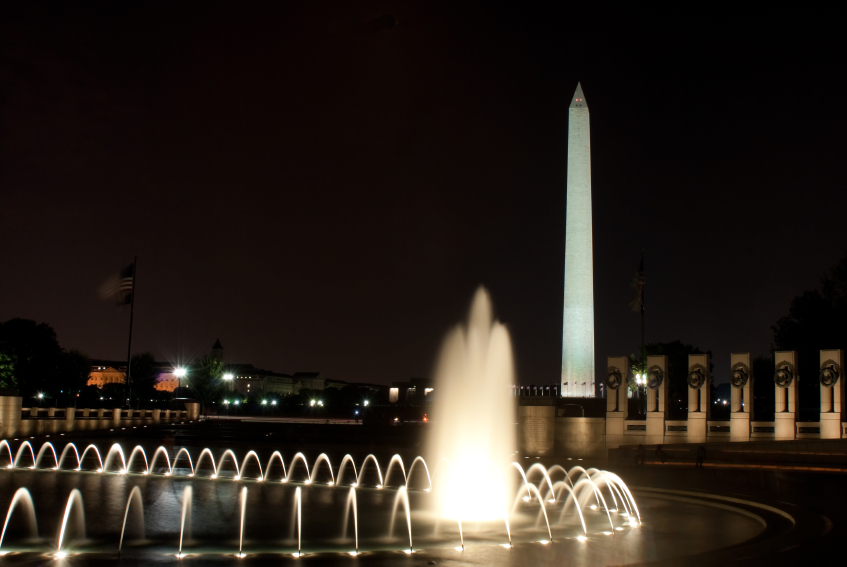 A nighttime view of the Washington Monument illuminated against the dark sky. In the foreground, a circular fountain with white streams of water is lit up, much like the aspirations of those pursuing Hill internships. The scene includes nearby flags and structures partially visible under the dim light.