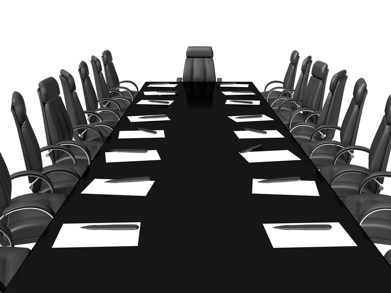 A long black conference table surrounded by black leather chairs evokes a sense of formality, as if awaiting a council meeting. Each seat has a pad of paper and a pen placed in front of it. The room is empty, and the chair at the head of the table is prominently positioned. The background is white.