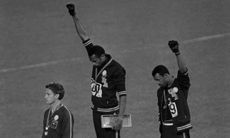 Black and white photo of three athletes standing on a podium. Two of them, displaying immense courage, have their fists raised and heads bowed, wearing black gloves. The third athlete stands with his arms at his sides, looking forward. All are dressed in athletic gear with medals on their necks.