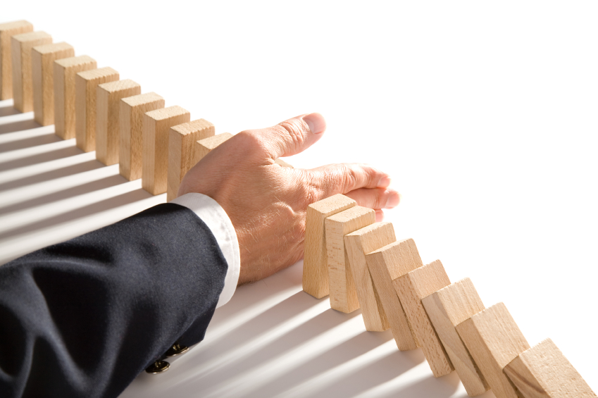 A hand wearing a suit sleeve stops a series of wooden dominoes from toppling in sequence. The dominoes are partially falling, but the hand intervenes to prevent the rest from tumbling over, expertly halting the chain reaction—a perfect metaphor for effective performance management.