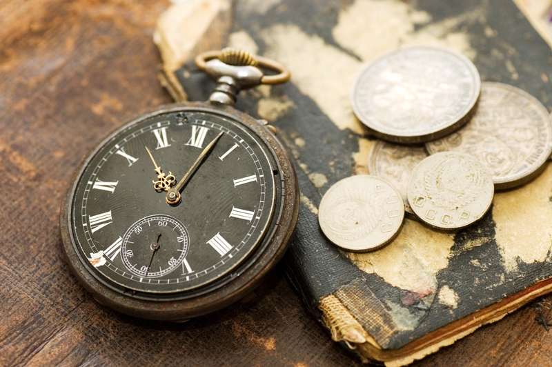 A vintage pocket watch with a dark face and Roman numerals lies on a weathered, old book with peeling edges. Several aged coins are scattered beside the watch, all resting on a rustic wooden surface, subtly reminding us of the timeless essence of productivity.