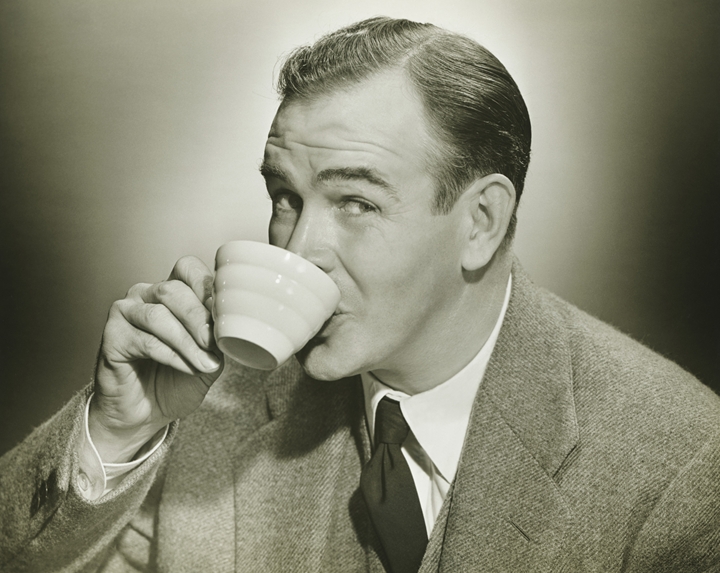 A vintage sepia-toned photograph of a man wearing a suit and tie, taking a break while sipping from a white striped teacup. He is smiling and looking towards the camera. The background is plain and slightly blurred, highlighting the subject.