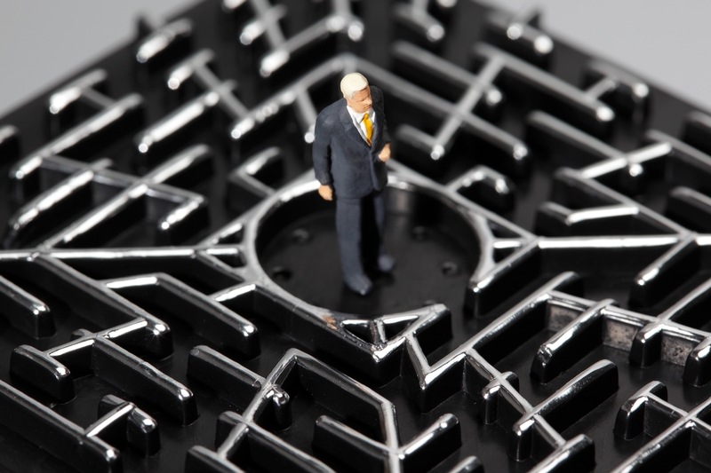 A miniature figurine of a man in a suit stands in the center of a black, metal maze with raised pathways. The man appears to be contemplating his job as he navigates the intricate labyrinth. The image highlights themes of complexity and challenge.