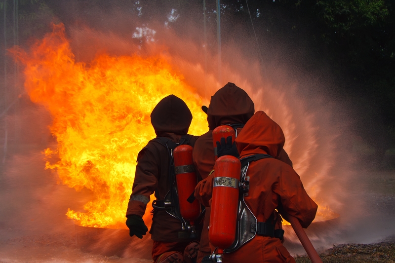 Three firefighters in brown protective gear and oxygen tanks work together to extinguish a large blaze with water hoses. The intense fire service effort creates bright orange flames and thick smoke, while the water spray forms a mist, partially obscuring the view.