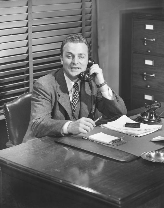 Black and white photo of a man in a suit sitting at a desk with a phone in his hand. He is smiling and looking forward, embodying the optimism of a job seeker. The desk is cluttered with papers, a notepad, and a pen. There are closed blinds behind him and a filing cabinet to his right.
