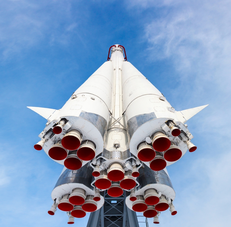 A low-angle shot of a white space rocket with multiple red exhaust nozzles, set against a blue sky with wispy white clouds. The image emphasizes the intricate details of the rocket's engines and structure, conveying a sense of power and technological advancement driven by precise engineering habits.