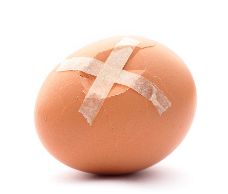 A brown egg with noticeable cracks is repaired with two pieces of transparent tape forming an X shape on a white background, symbolizing the delicate balance often required in one's career.